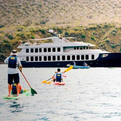Travelers paddleboarding and kayaking in front of a small Galapagos expedition yacht anchored by an arid island